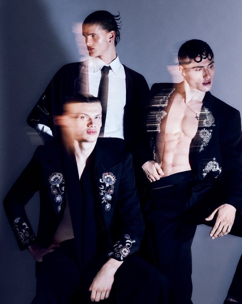 Three young adult men pose in dark suits with floral embellishments against a grey backdrop in a fashion editorial style shot.