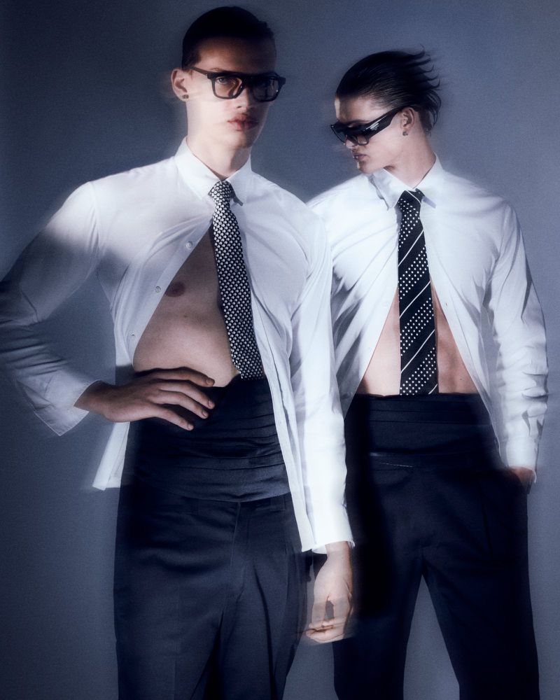 Two adult men pose indoors wearing white shirts, dark pants, ties, and sunglasses. One shirt is unbuttoned, revealing his chest.
