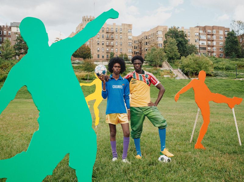 Two dark-skinned men pose in soccer uniforms on a grassy field, surrounded by painted soccer player silhouettes, with apartments in background.