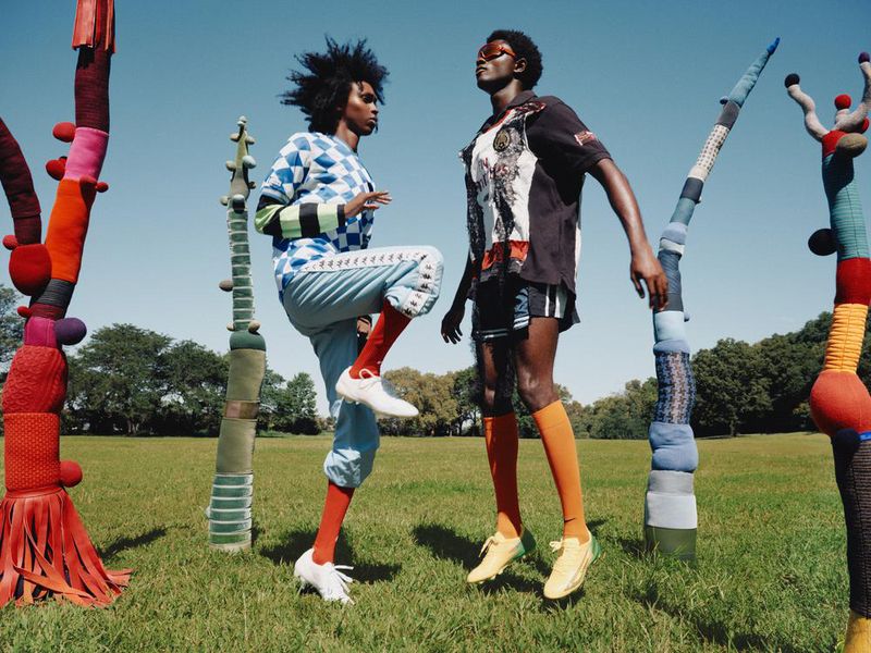 Two slender figures strike poses on a grassy field adorned with tall, whimsical, fabric-covered sculptural forms under a bright blue sky.