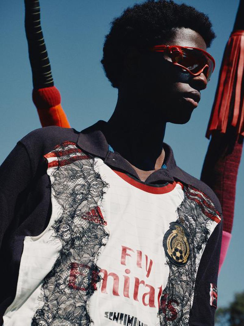 Fashionable black male model with red sunglasses poses wearing a white soccer jersey, with blue sky background and fringe accents.