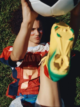 Person lies on grass with soccer ball, wearing face paint, soccer jersey, lace undershirt, and yellow soccer cleat, looking determined.