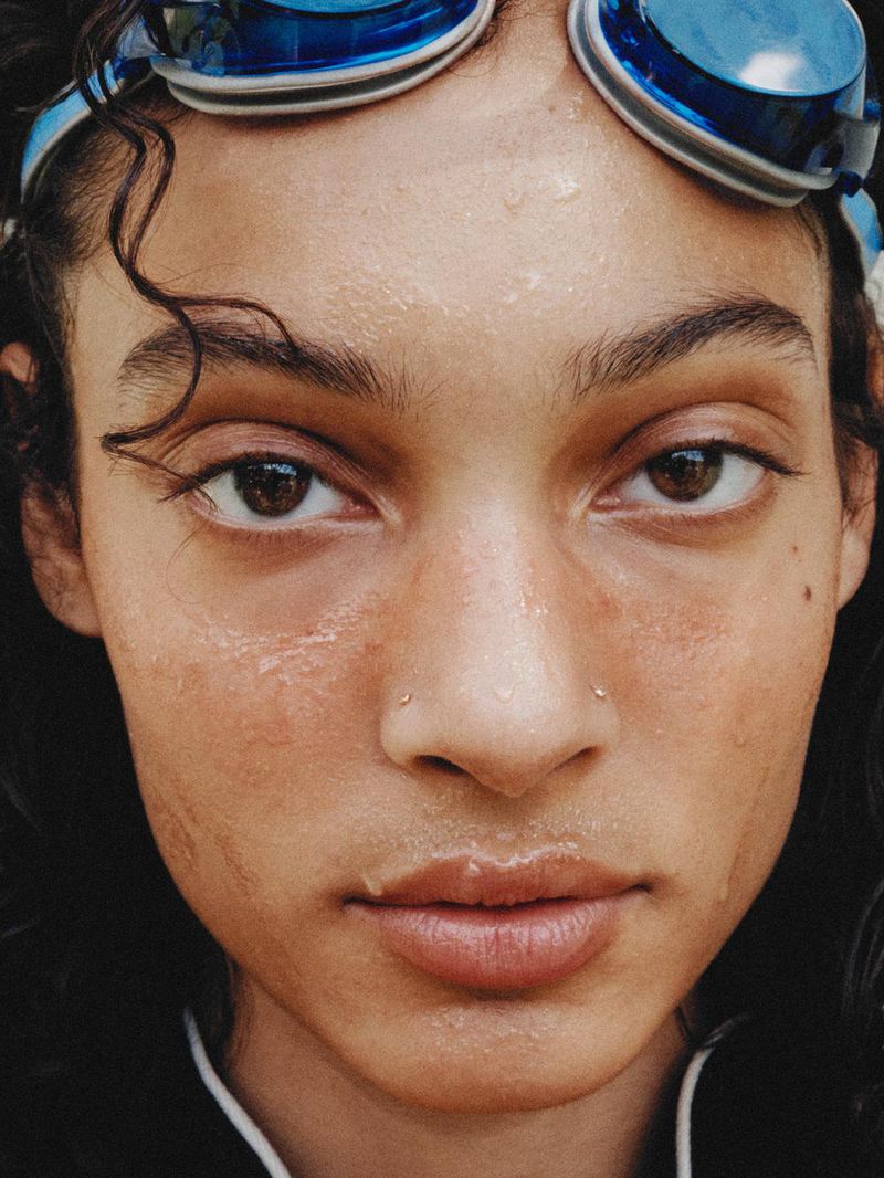 Close-up portrait of a young adult woman with wet skin, wearing swimming goggles on her forehead, and featuring natural, glossy lips.