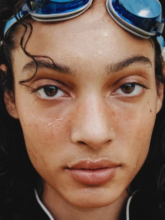 Close-up portrait of a young adult woman with wet skin, wearing swimming goggles on her forehead, and featuring natural, glossy lips.
