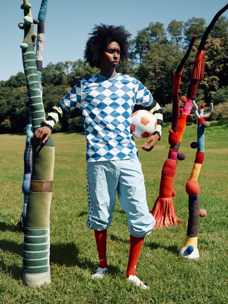 A man in a blue and white diamond-patterned soccer jersey holds a soccer ball in a grassy field with abstract sculptural elements.