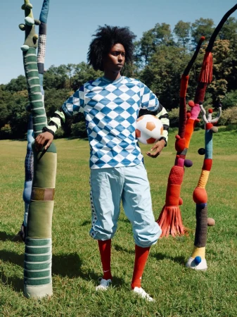A man in a blue and white diamond-patterned soccer jersey holds a soccer ball in a grassy field with abstract sculptural elements.