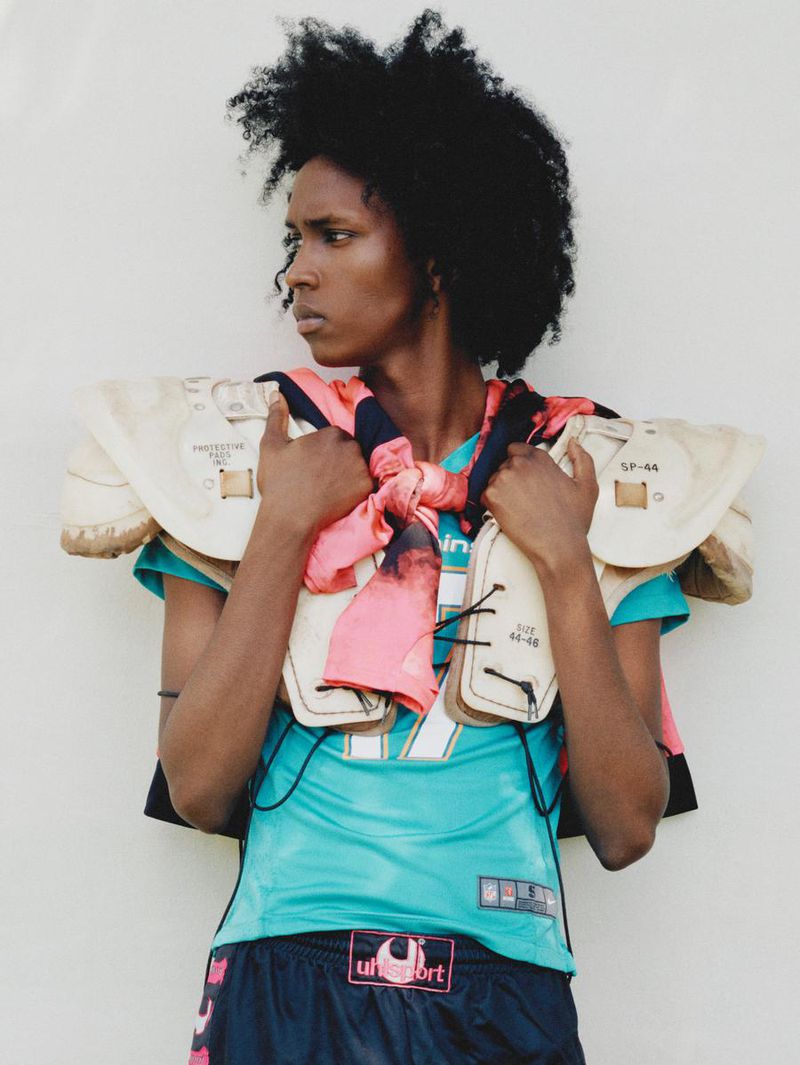 Person wearing sports gear and football pads against a white wall, turquoise jersey and scarf, stylish and edgy.