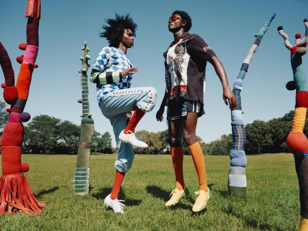 Two athletic models posing outdoors in a park surrounded by tall whimsical yarn-wrapped sculptures under a clear sky.