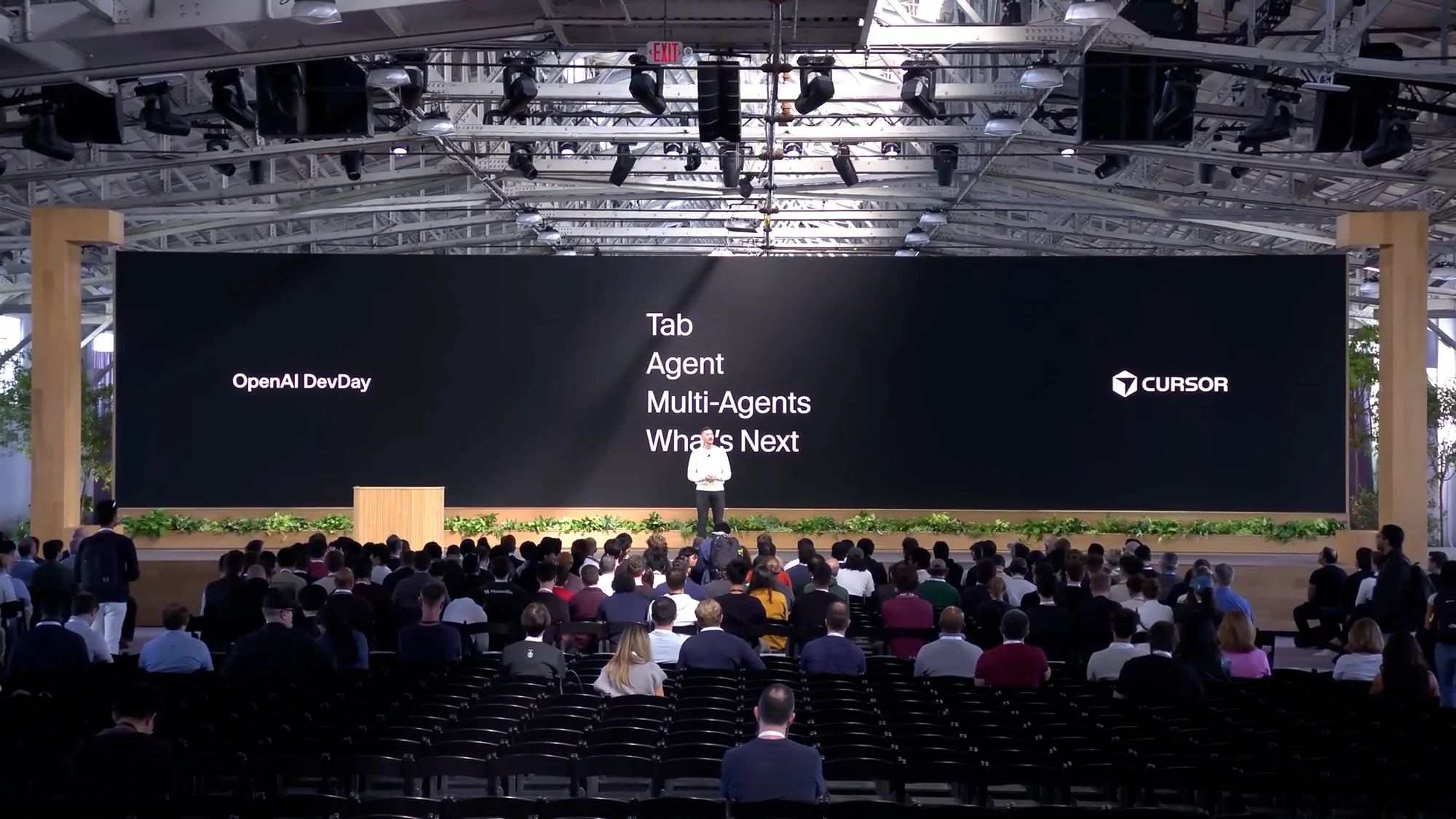 Audience at OpenAI DevDay event watches a speaker on stage in a large indoor space with a large screen displaying the event's theme.