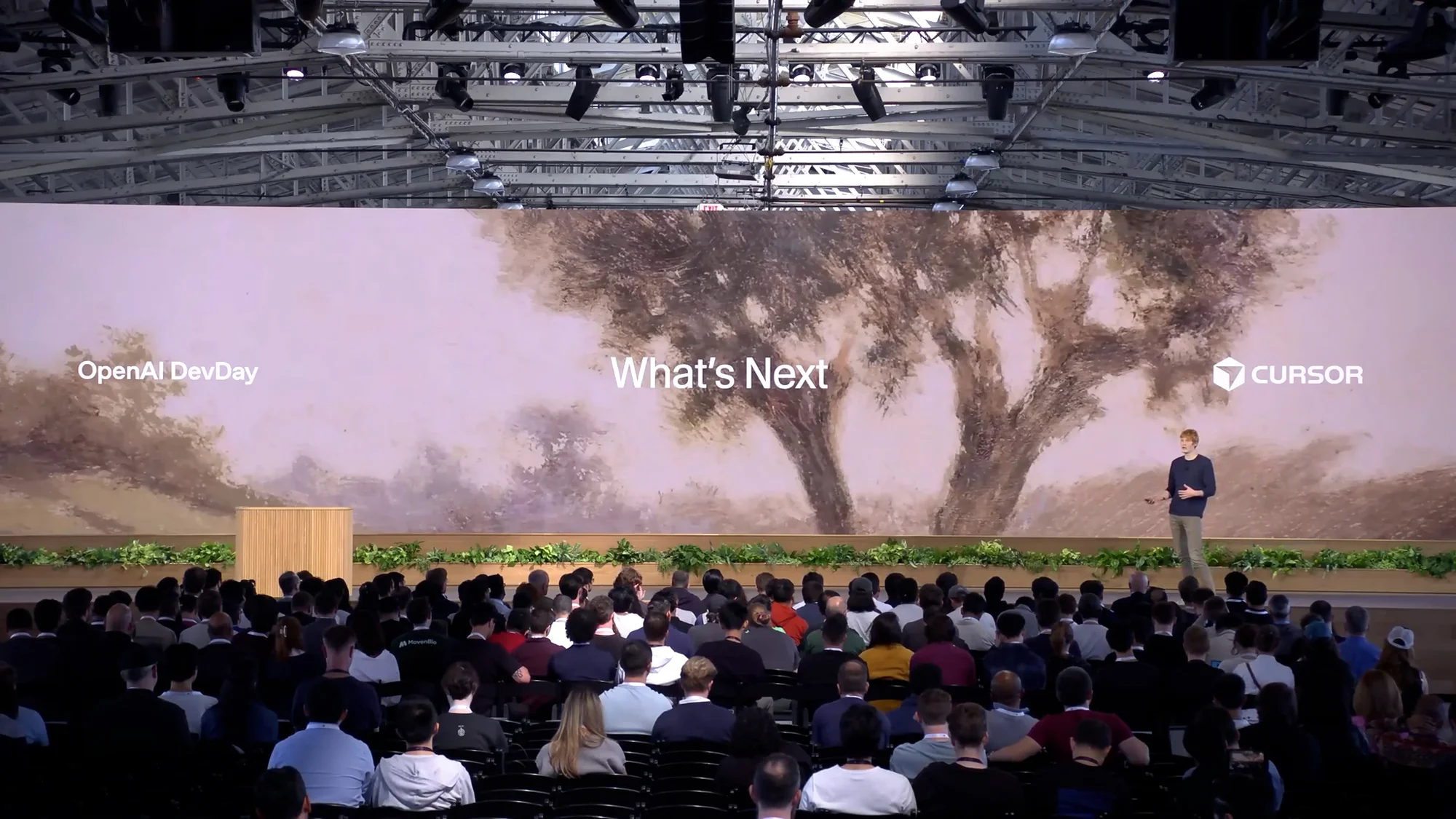Audience watching a speaker at a tech conference with a screen displaying 'What's Next' and the Cursor logo on stage.