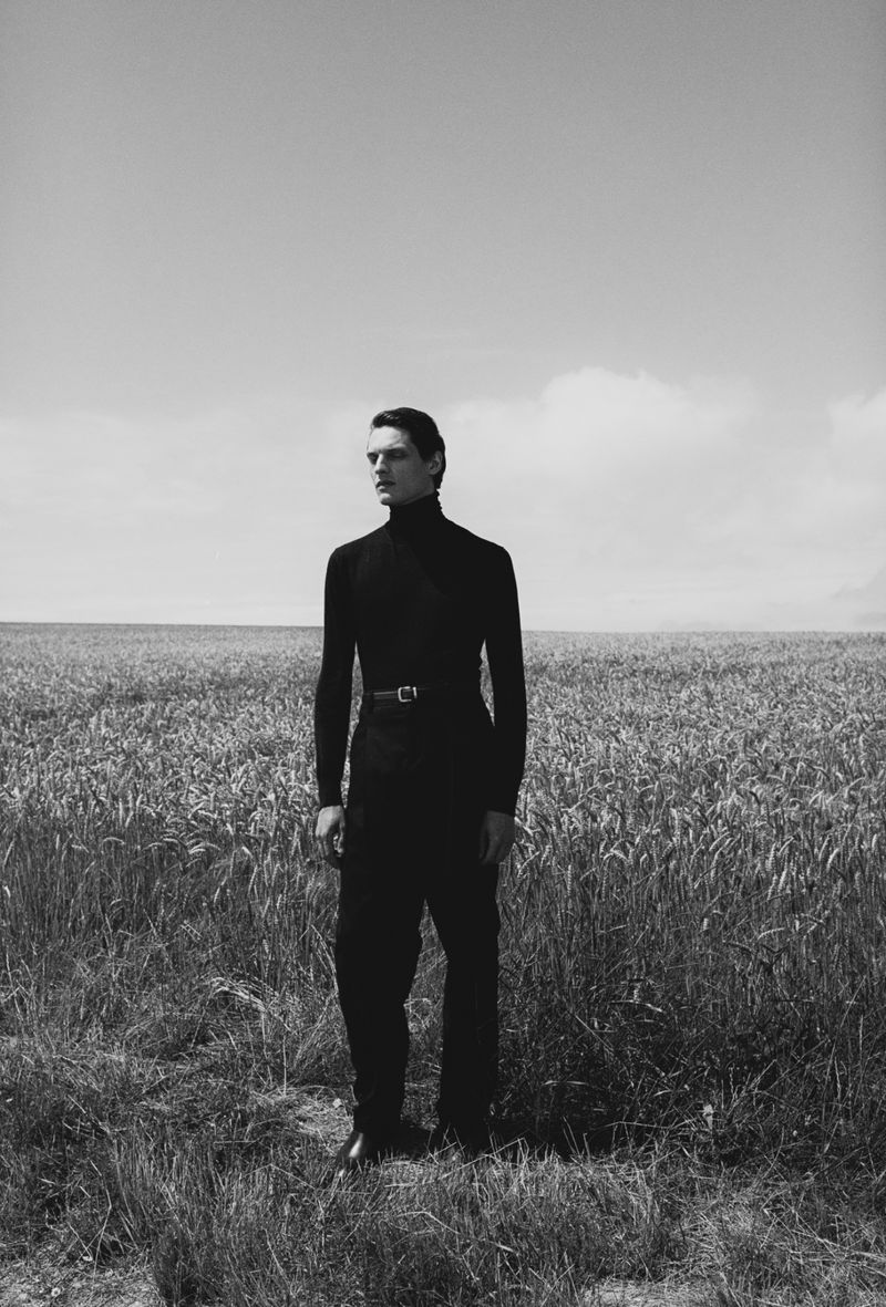 Monochrome portrait of a slender man in black turtleneck standing in field of wheat under a cloudy sky looking off into the distance.