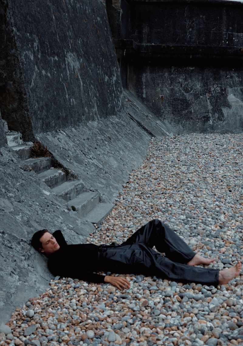 Adult male laying on a pebble beach near a concrete wall, wearing a black long sleeve shirt and shiny black pants, barefoot.