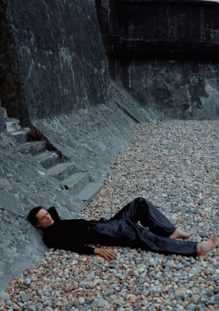 Adult male laying on a pebble beach near a concrete wall, wearing a black long sleeve shirt and shiny black pants, barefoot.