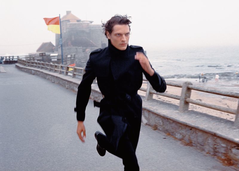 Male model in a long black coat running along a coastal path near a stone fortress with the sea behind him.