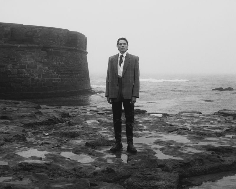 Monochrome shot of a man in a suit standing on a rocky shore with the ocean in the background near an old stone structure.