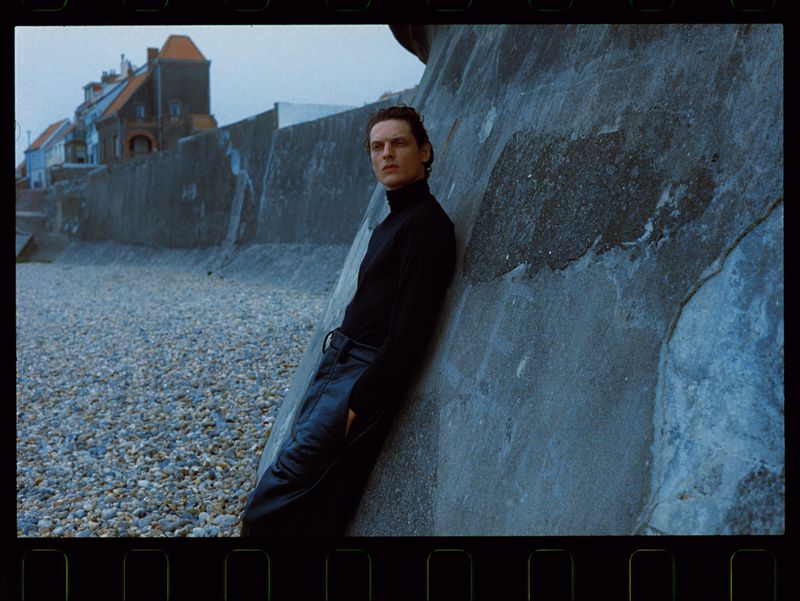A lone male figure leans against a weathered wall on a rocky beach, wearing a black turtleneck and black leather pants, under an overcast sky.