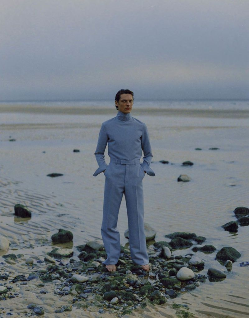 Fashion shot of a young adult male in a light blue jumpsuit, standing barefoot on a rocky beach with the ocean and sky in the background.