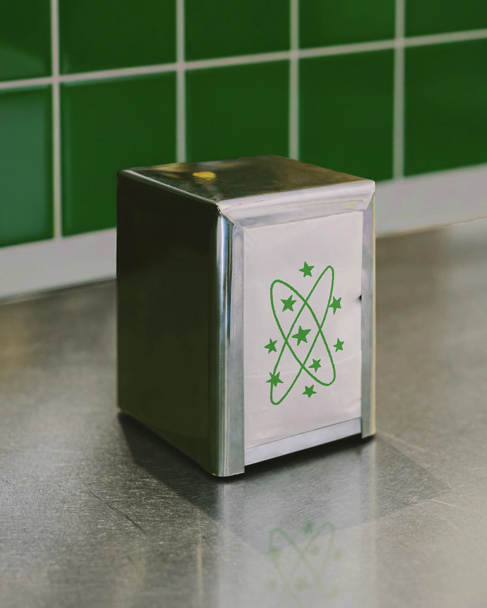 Metal napkin holder with a stylized atom design in green on a countertop with green tiled backsplash, minimalist studio shot.