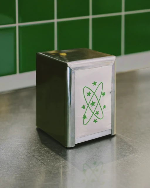 Metal napkin holder with a stylized atom design in green on a countertop with green tiled backsplash, minimalist studio shot.