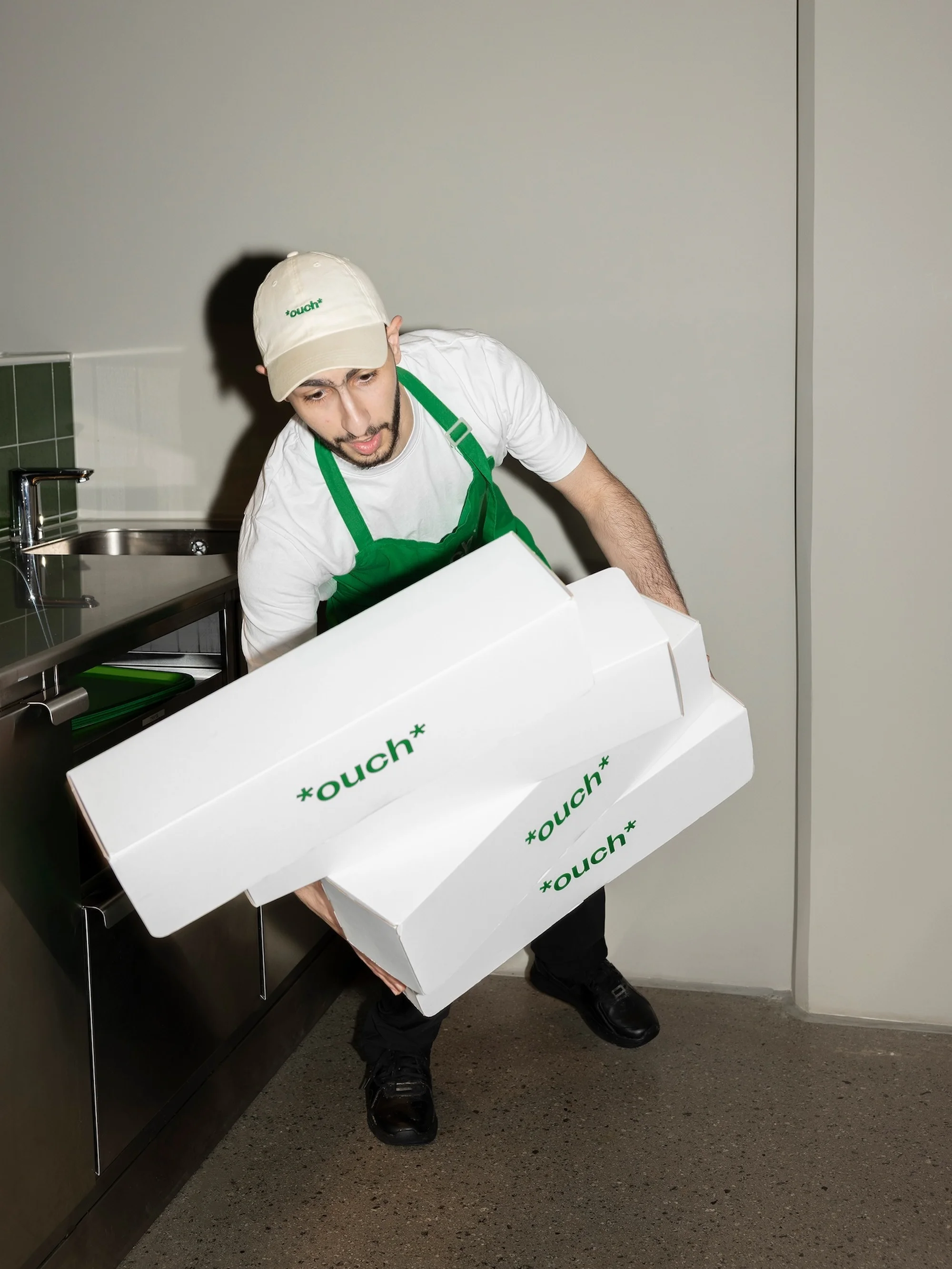 Man in green apron carries stack of white boxes marked 'ouch' in a commercial kitchen with stainless steel sink and neutral wall.
