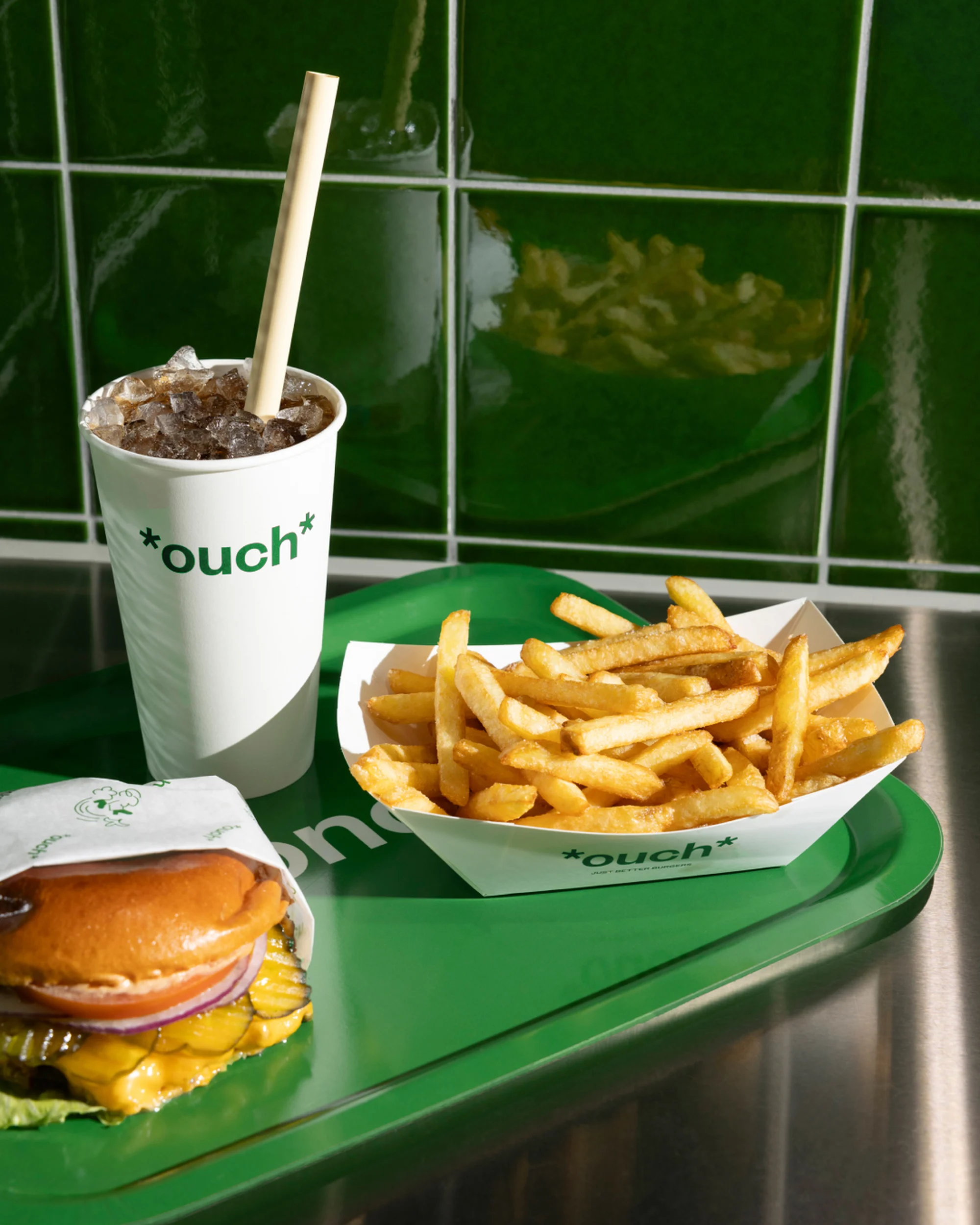 Overhead shot of a burger, fries, and drink on a green tray against a green tile wall. The meal is lit by natural, diffused light.
