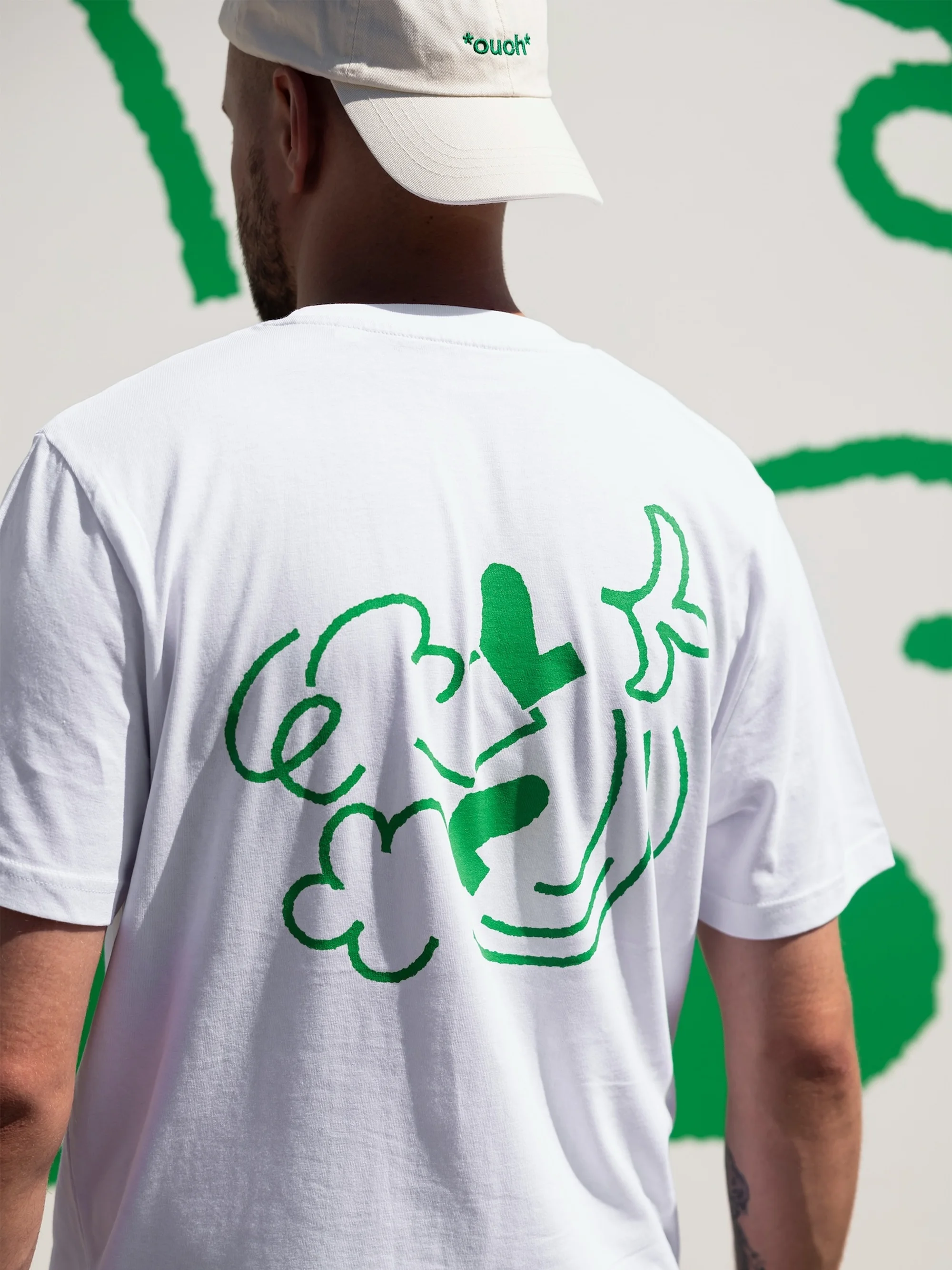 Man in white t-shirt with green cloud design, wearing a beige baseball cap, stands in front of a white wall with green design.