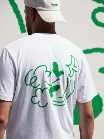 Man in white t-shirt with green cloud design, wearing a beige baseball cap, stands in front of a white wall with green design.