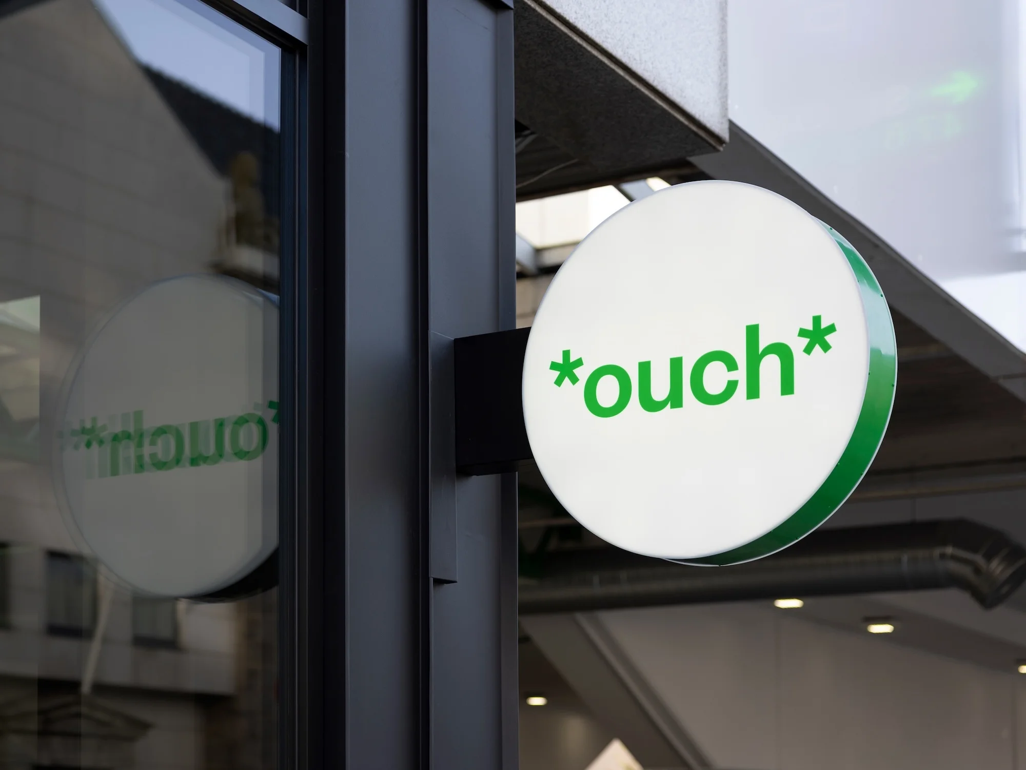 Illuminated round sign with the word '*ouch*' in green, mounted on a building's exterior, reflecting the surroundings in a nearby window.