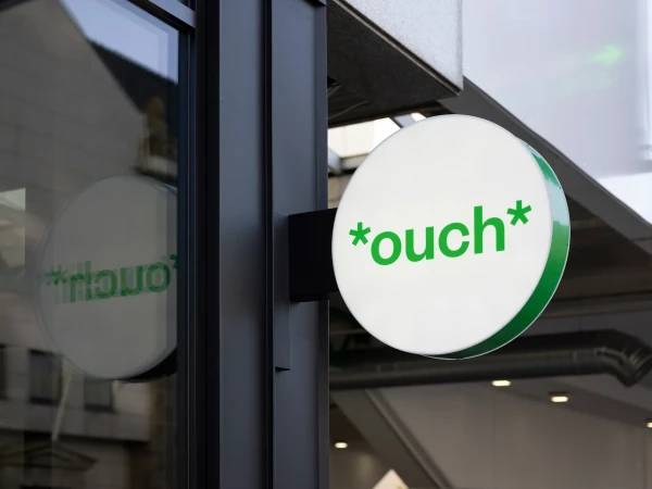 Illuminated round sign with the word '*ouch*' in green, mounted on a building's exterior, reflecting the surroundings in a nearby window.