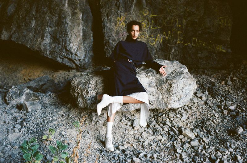A fair-skinned adult in a black turtleneck and skirt, white boots, and socks sits on a large rock inside a cave with graffiti.