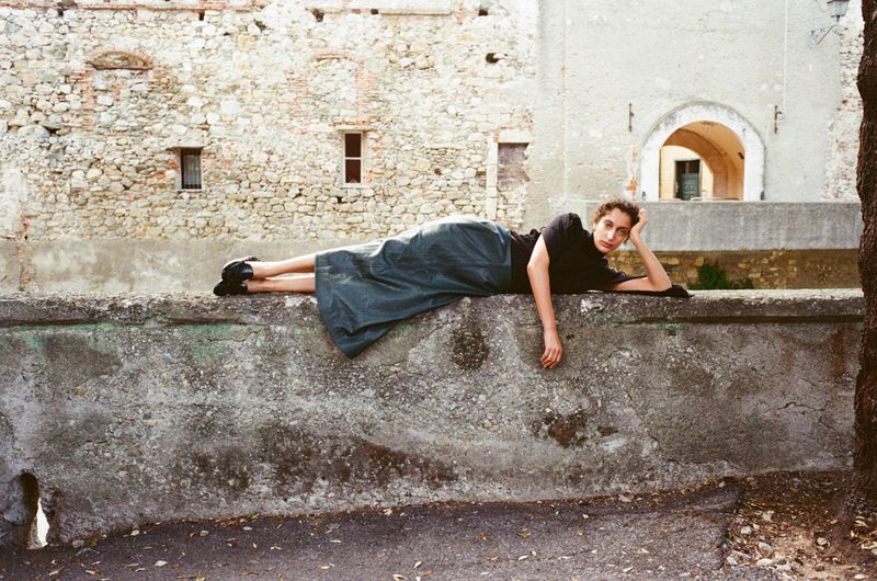 Woman in a dark top and long green skirt laying on a stone wall outside a stone building with arched doorway in the background.