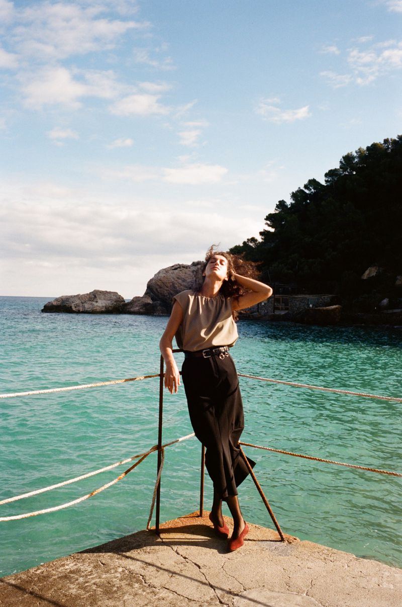A solo female figure in stylish attire stands on a concrete pier by the sea, enjoying sunlight in a fashion or lifestyle context.