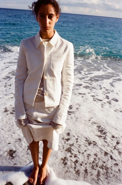 Woman in a linen skirt suit standing barefoot in the surf with ocean and sky in the background, creating a calm, natural fashion aesthetic.