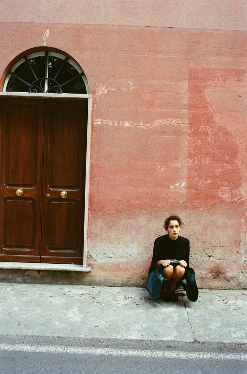 Adult female with dark hair sits on the sidewalk in front of a textured salmon-colored building with a brown door with an arched window.