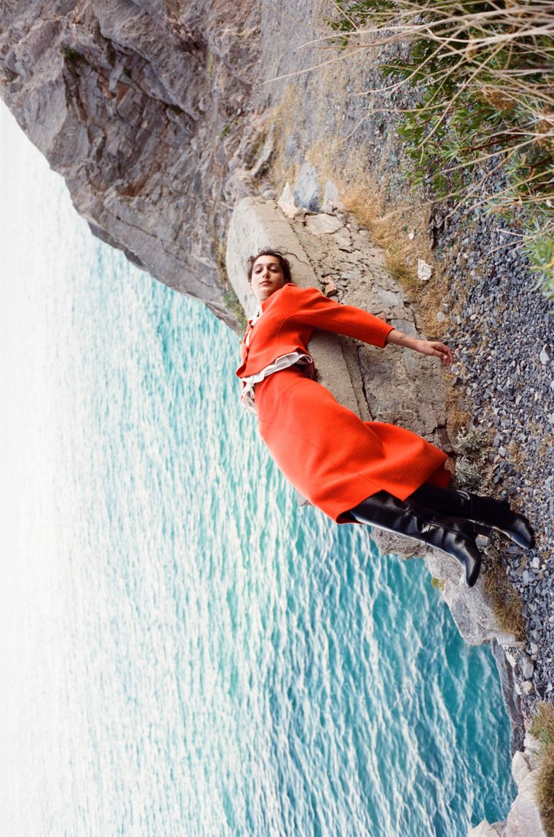 A woman in an orange skirt suit and black boots lies on a concrete ledge near a rocky cliff overlooking the ocean.
