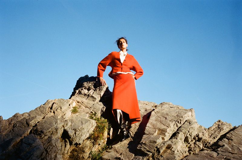 Fashionable woman in orange skirt suit poses atop rocky mountain against clear blue sky, showcasing outdoor editorial style.