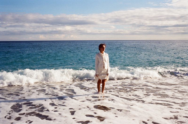 Woman in a white coat stands in the ocean as waves crash. The background shows a cloudy sky meeting the turquoise water.