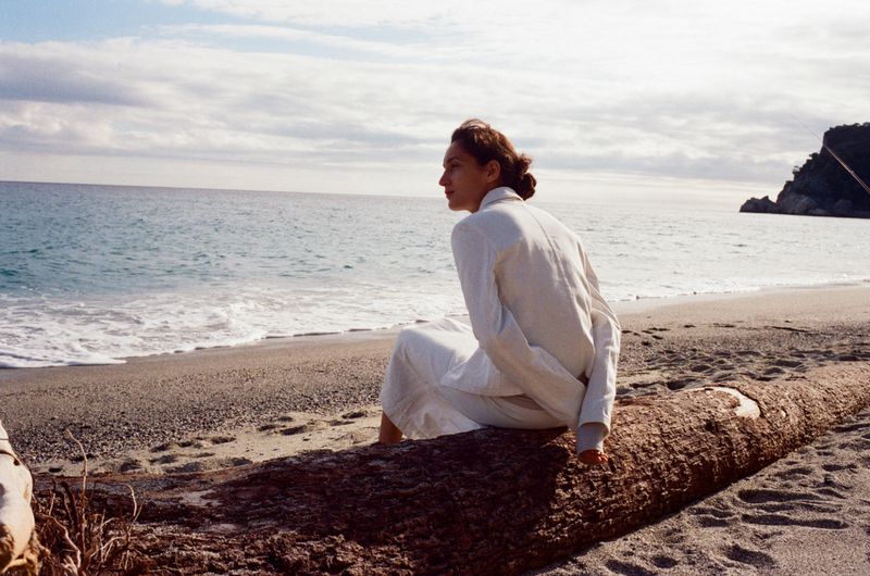 Woman in white sitting on a log at a beach, looking at the ocean with cloudy sky, capturing a serene moment in nature.