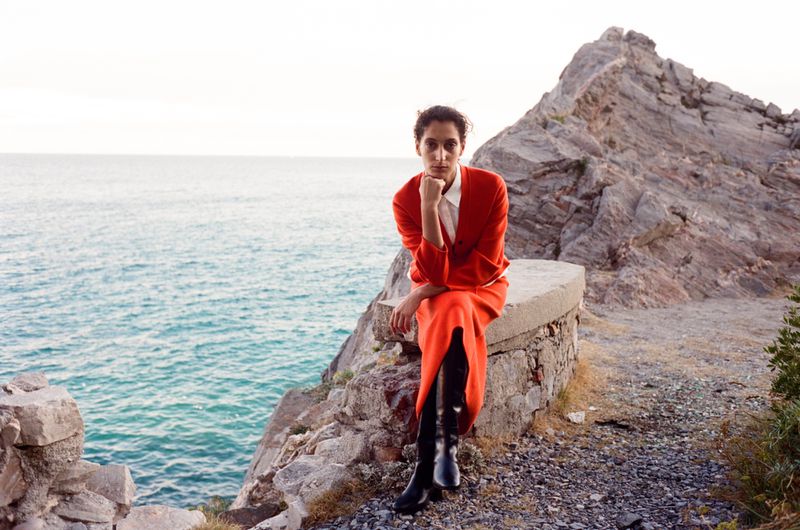 A young adult woman in a red dress and boots sits on a stone ledge overlooking the ocean, with a rocky cliff behind her.