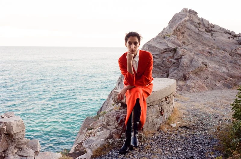 A young adult woman in a red dress and boots sits on a stone ledge overlooking the ocean, with a rocky cliff behind her.