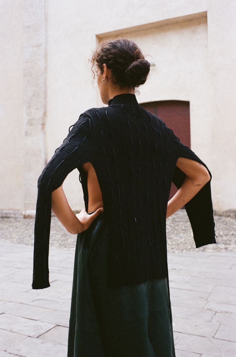 Back view of a female fashion model wearing a black knit top and dark green skirt in front of a light-colored stucco building.