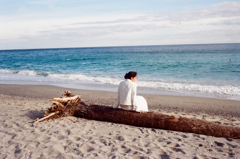 Woman in white sitting on driftwood on a sandy beach with turquoise water and a partly cloudy sky in a tranquil outdoor setting.