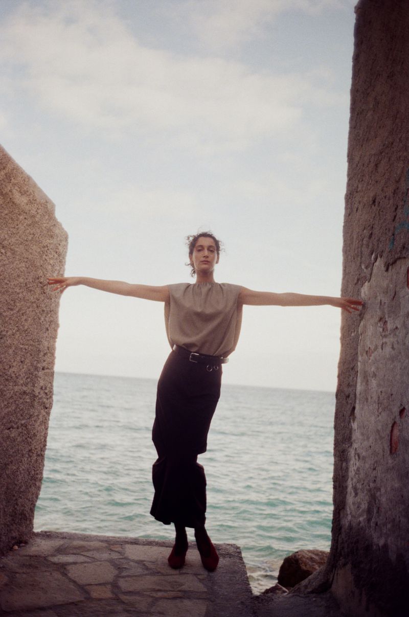 A woman in a beige top and black skirt stands between stone walls with her arms outstretched at the ocean's edge.