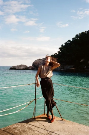 Model standing on a concrete pier overlooking turquoise ocean water with a rocky coastline under a blue sky.