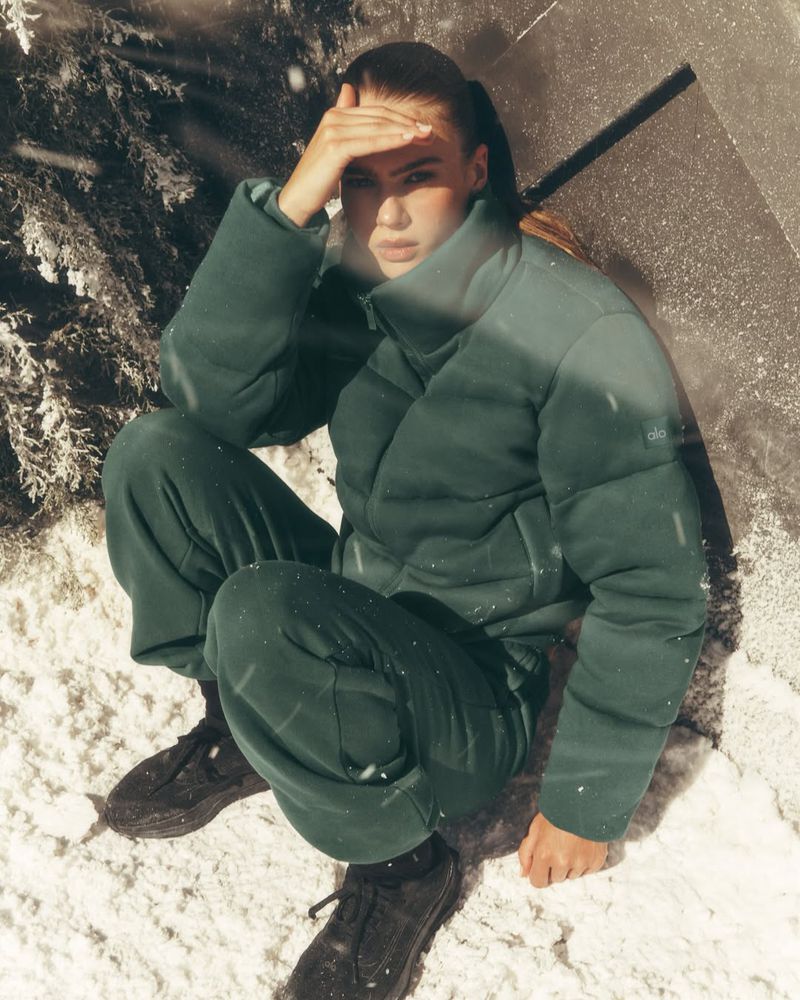 Brunette woman in a green puffy jacket and pants sitting in a snowy environment with a hand shielding her eyes from the light.
