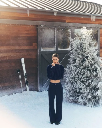 A young woman in dark workout clothes stands in the snow in front of a rustic wood building and a snow-covered evergreen tree.