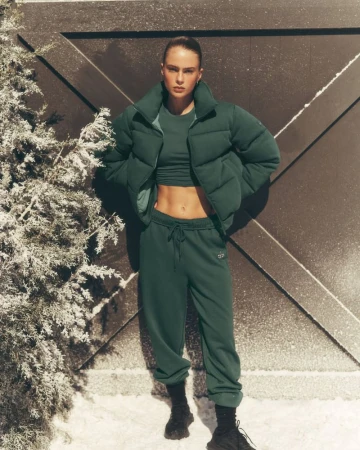 Athletic woman in sportswear: green crop top and joggers, puffer jacket, and sneakers. Posing outside a dark wood door in a snowy winter setting.