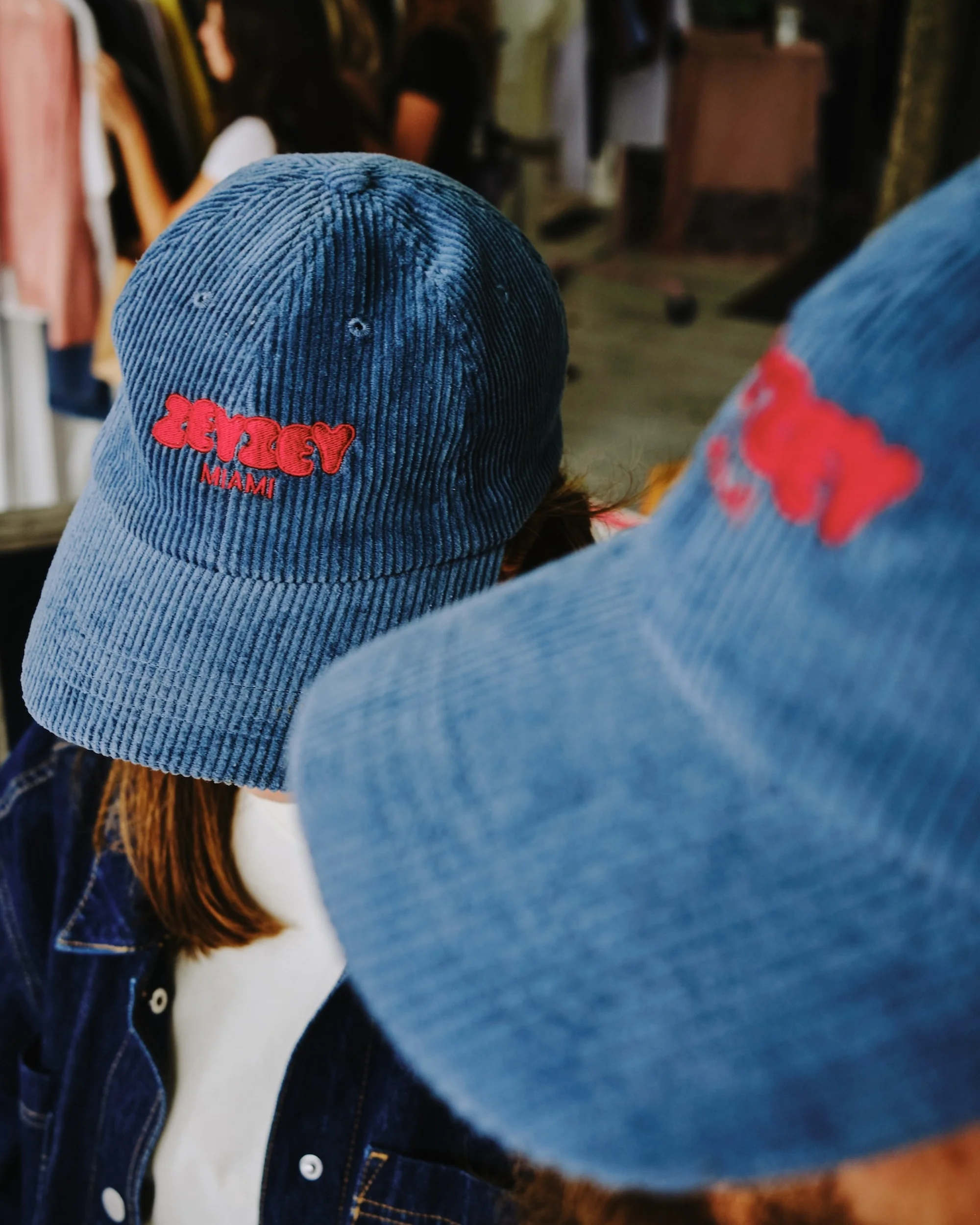 Two corduroy baseball caps with red lettering, one on a person and the other in the foreground, with clothing rack background.