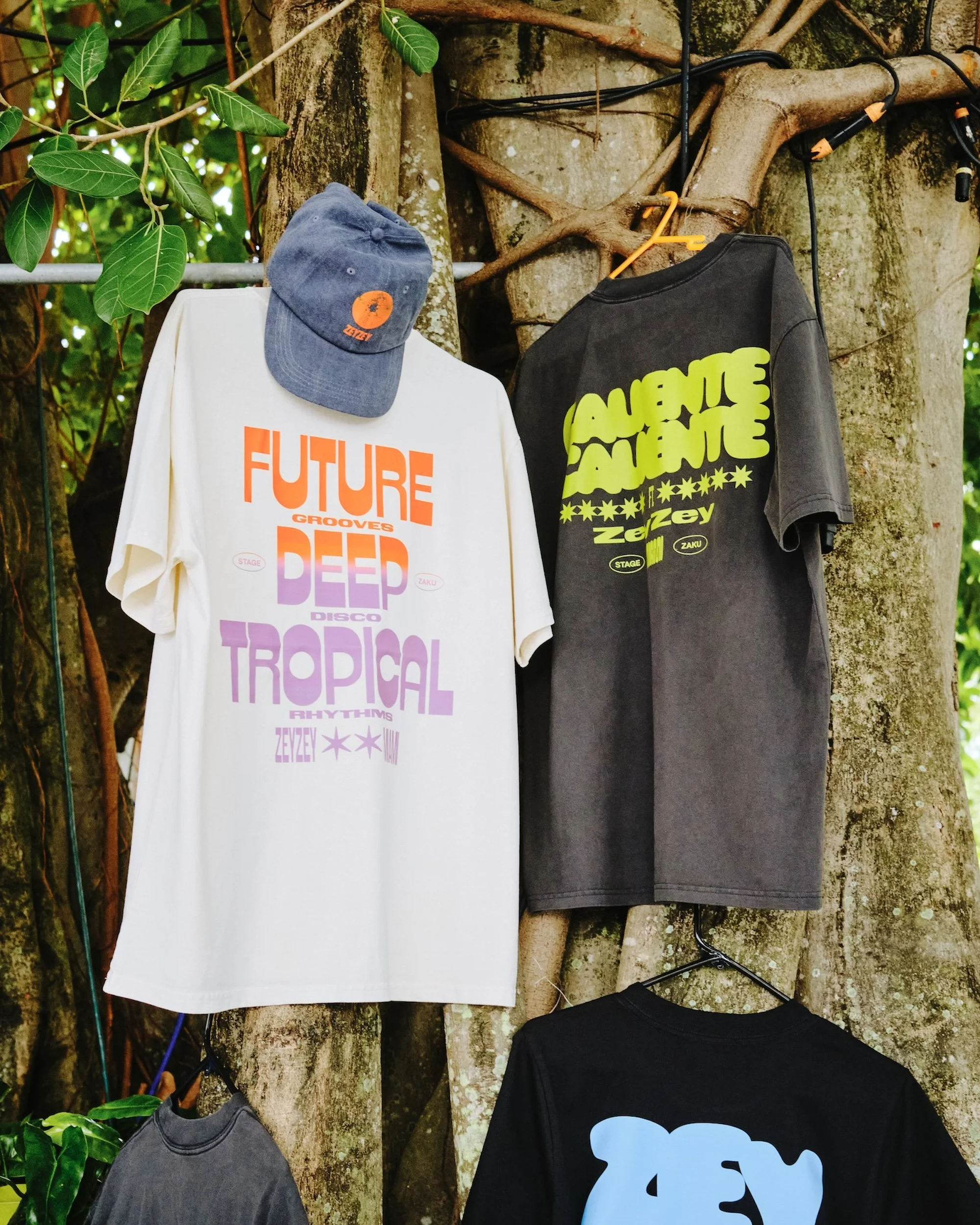 T-shirts and a cap are hung from a tree, creating an outdoor display of apparel with colorful text designs on the fabric.