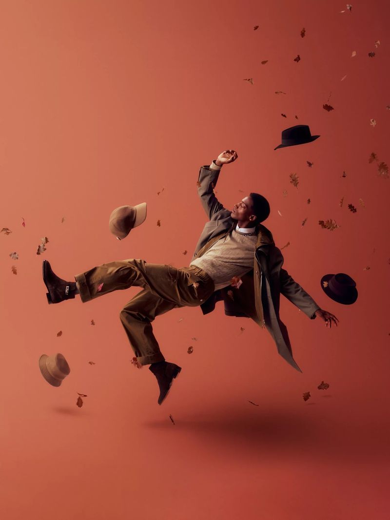 Stylish Black man in gray coat levitates mid-air against red backdrop amidst falling leaves and hats for fashion editorial, studio lighting.